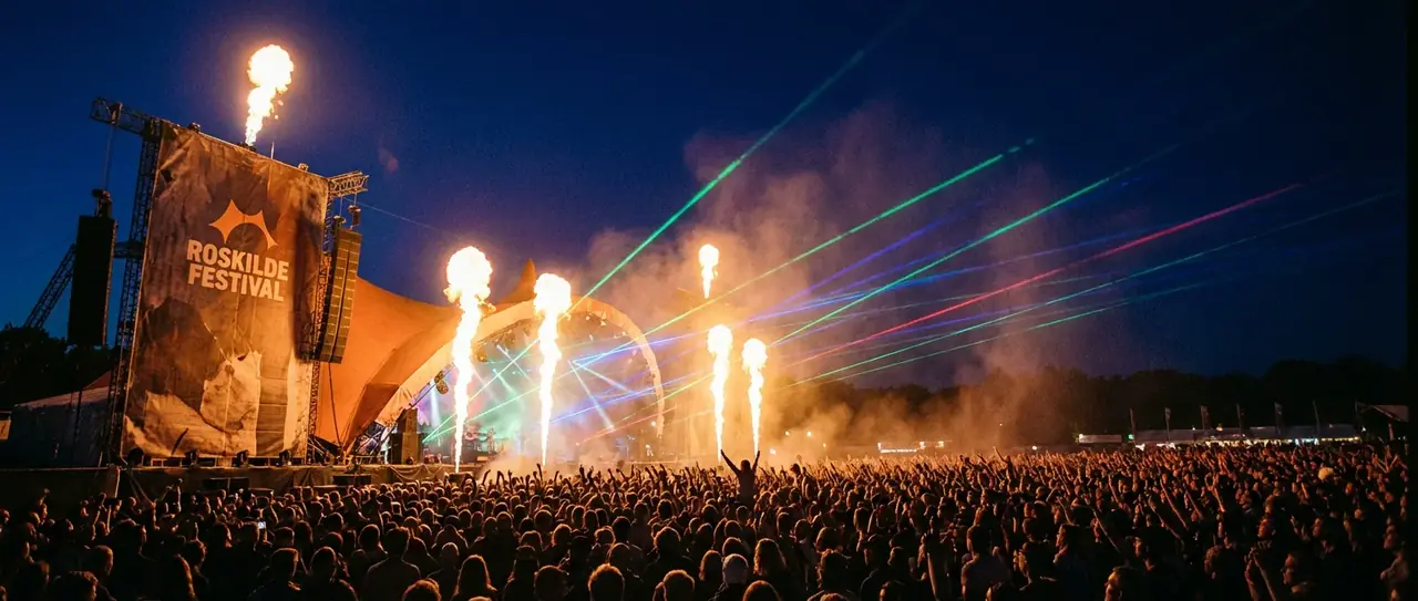 Publikum med hænderne i vejret foran en oplyst festivalscene om aftenen.
